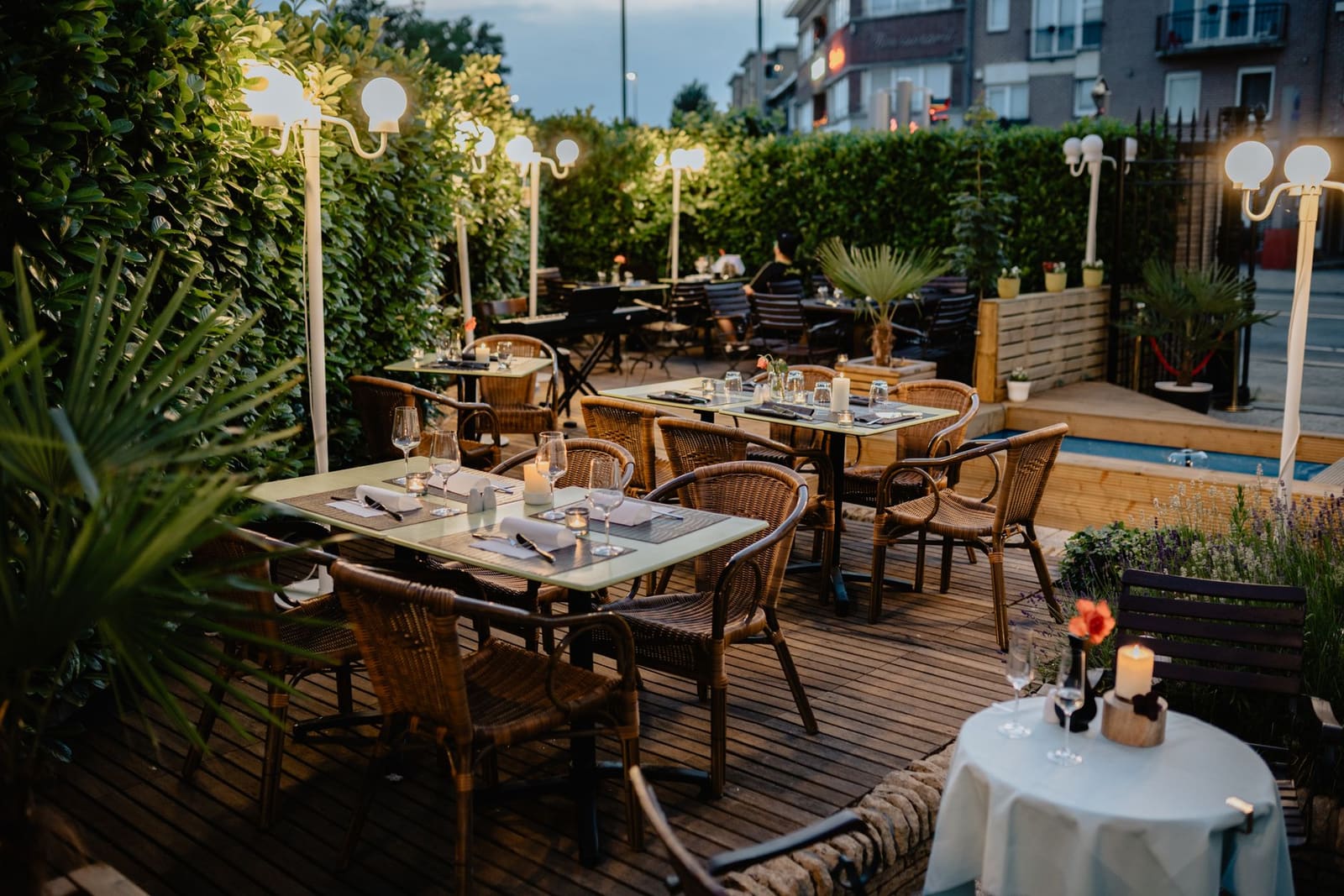Terras met gedekte tafels en witte bollampen langs een haag in de schemering